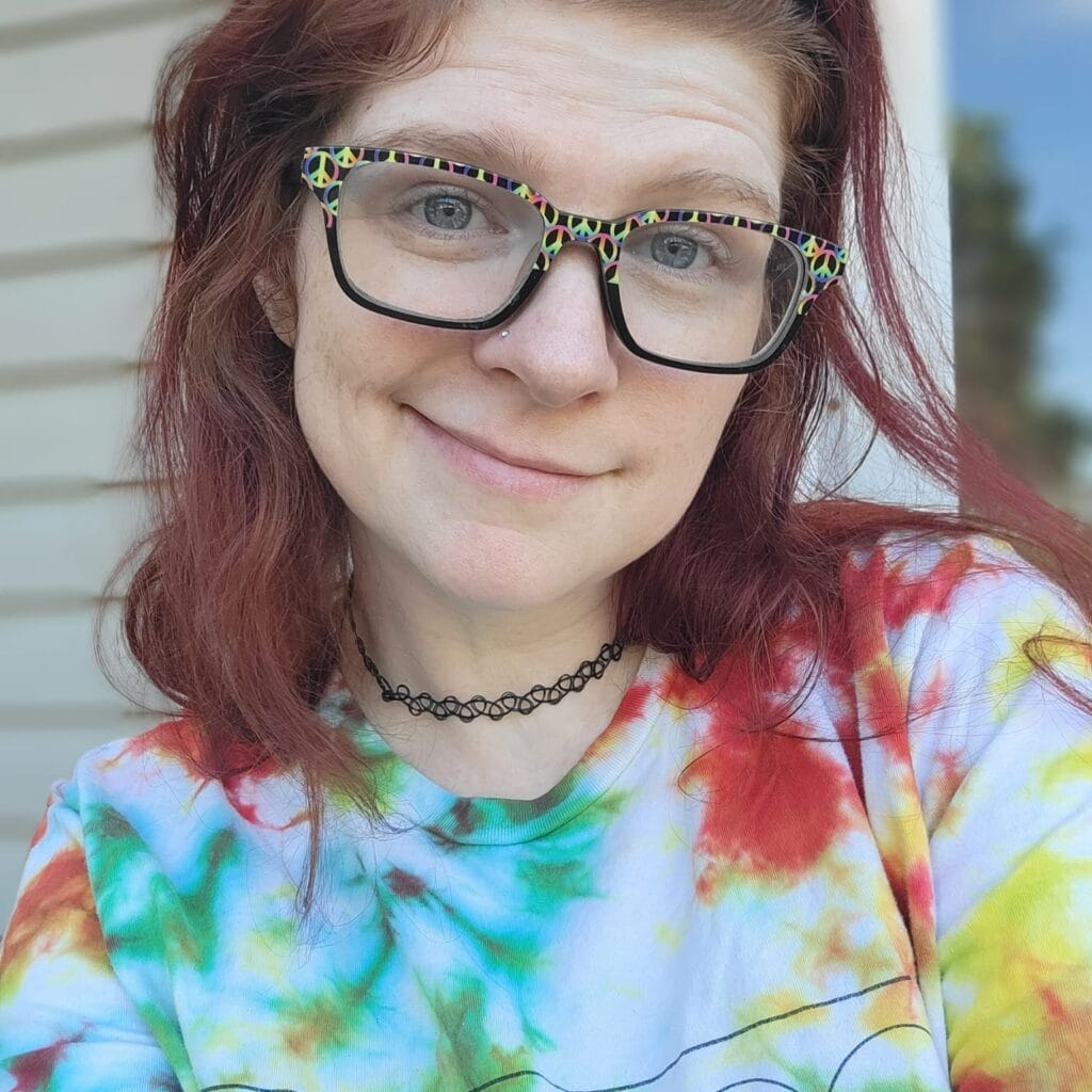 Smiling woman with red hair and colorful peace-sign glasses wearing a tie-dye shirt and choker necklace, radiating warmth and authenticity in a casual outdoor setting